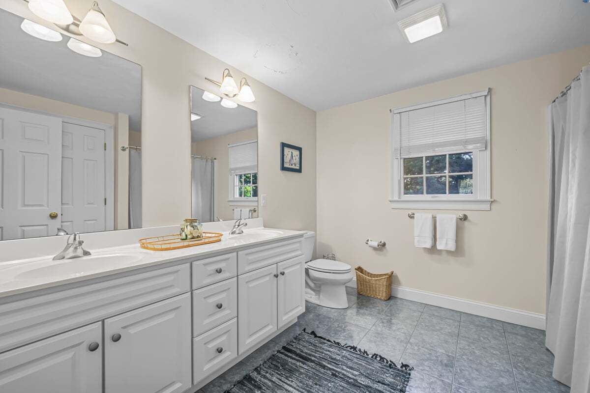 A bathroom with a white vanity, a large mirror, a toilet, and a window with blinds.