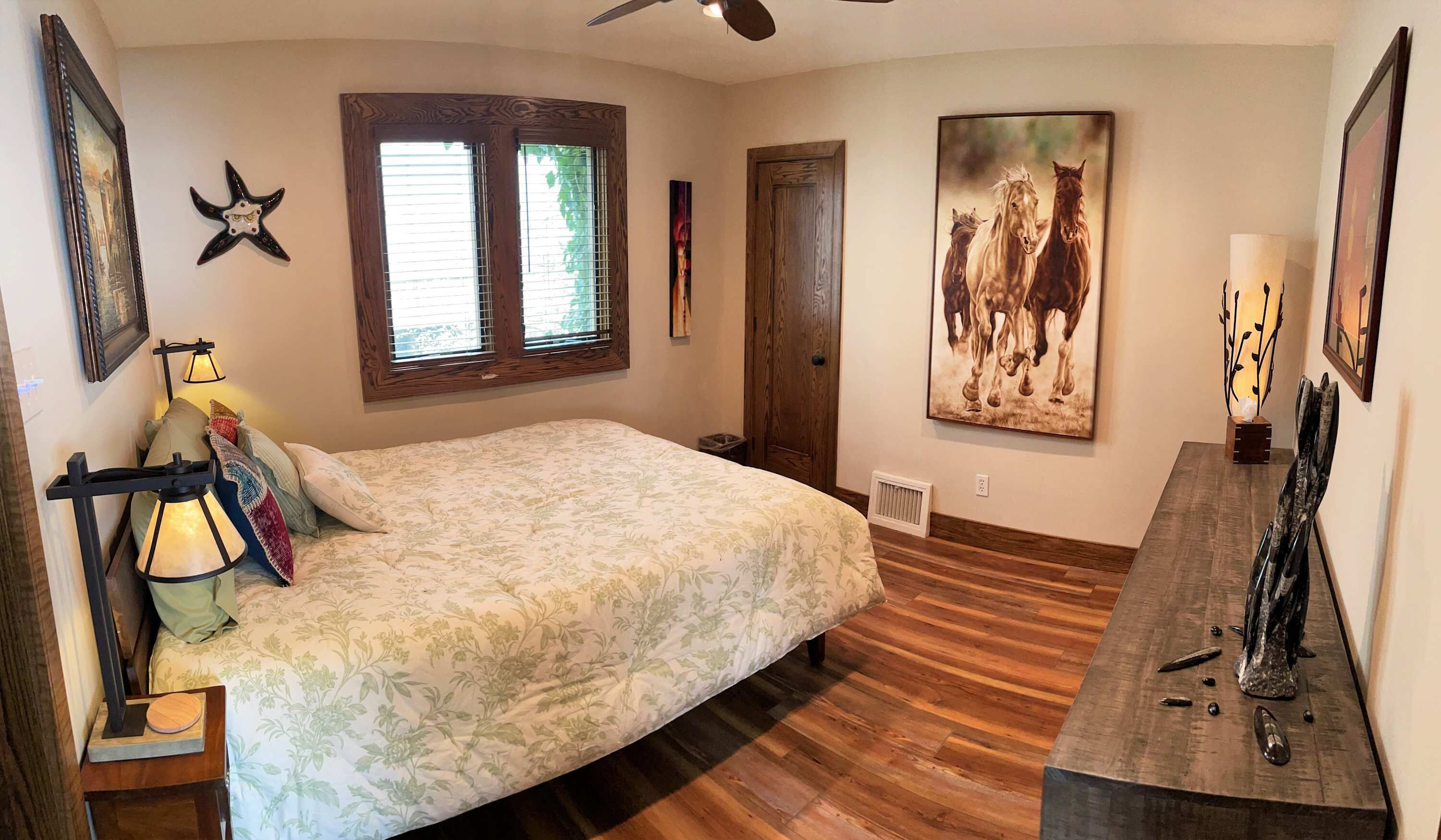 A bedroom with a bed, wooden flooring, a ceiling fan, a window with blinds, and a door.