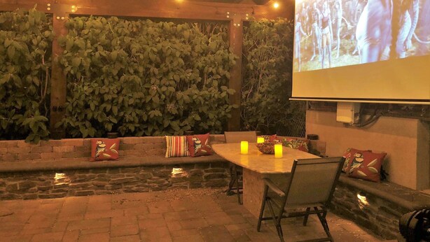 Patio esterno con una panchina in pietra, un tavolo con candele e uno schermo di proiezione che mostra una scena.