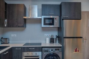 A modern kitchen with dark wood cabinets, stainless steel appliances including a microwave, oven, and refrigerator, and a built-in washing machine. - Loxwood House by Cozy (Accra)