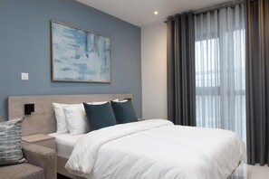 A modern hotel room with a large bed, a headboard, a bedside table, and a nightstand. There is a window with curtains, a wall-mounted light, and a painting on the wall. - Loxwood House by Cozy (Accra)