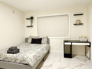 Un dormitorio con cama, un escritorio, una ventana con persianas y dos estantes en la pared.