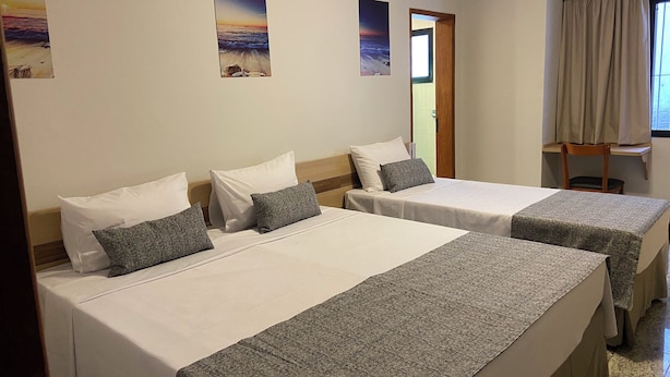 Habitación de hotel con dos camas, cada una con sábanas blancas y cojines grises. Hay dos cuadros en la pared, uno con una playa y el otro con una vista urbana. Se ve una ventana con cortinas color beige y una silla de madera junto a ella.
