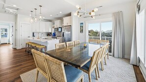 A modern kitchen with white cabinets, a central island, and a dining area with a large table and wicker chairs. - Summer Moon | 1,615 ft to the beach | Private Pool, Hot Tub, and Dog Friendly (Corolla)