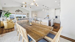A spacious living area with a large wooden dining table, wicker furniture, and a modern kitchen in the background. - Summer Moon | 1,615 ft to the beach | Private Pool, Hot Tub, and Dog Friendly (Corolla)