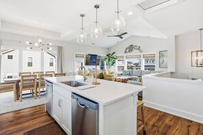 Eine moderne Küche mit Kochinsel, weißen Schränken und einem Geschirrspüler. Es gibt Pendelleuchten und einen Deckenventilator.