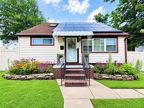 A single-story house with a solar panel roof, a front porch with steps, and a front yard with flowers and a stone border. - Spacious, Charming 4BR, 2BA Glen Burnie Home, Sleeps 9 (Glen Burnie)