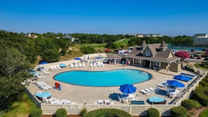 Une grande piscine entourée de chaises longues et de parasols, elle-même bordée d’une terrasse en bois et d’un bâtiment doté d’un toit à deux versants.