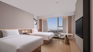 Quarto de hotel moderno com duas camas, uma mesinha, uma cadeira, televisão e uma janela grande com vista para a cidade.