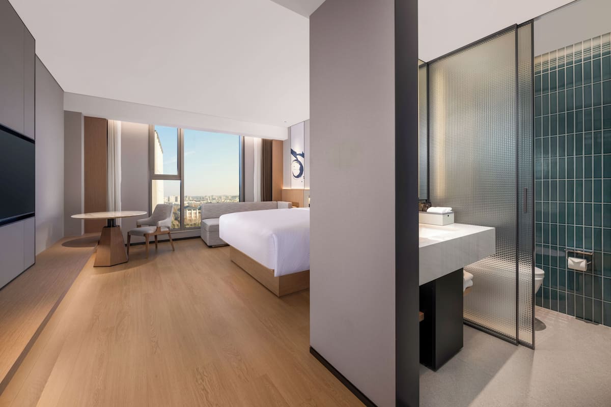 A modern hotel room with a large bed, a desk, a chair, a large window, and a bathroom with a glass partition.