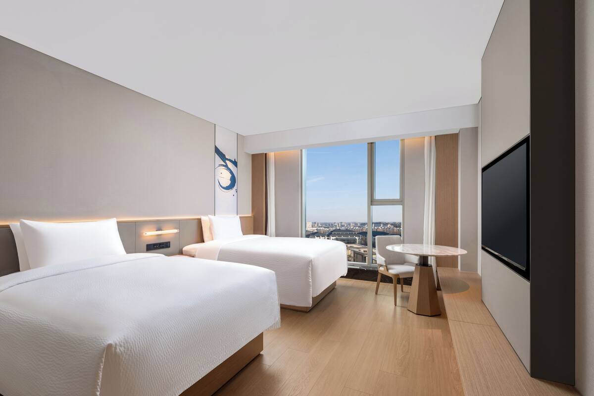 A modern hotel room with two beds, a small table, a TV, and a city view.