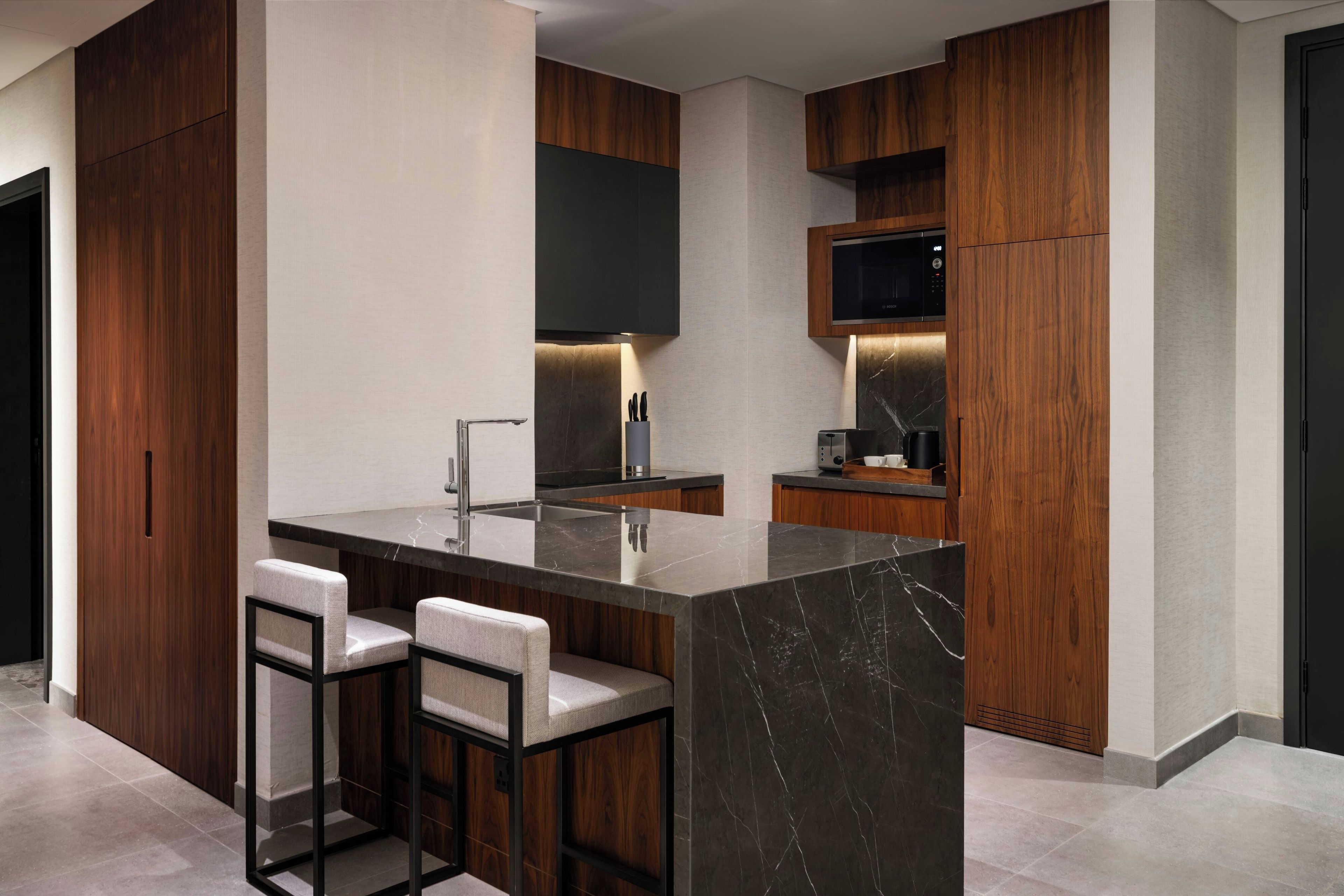 A modern kitchen with a marble island, bar stools, wooden cabinetry, and a built-in microwave.