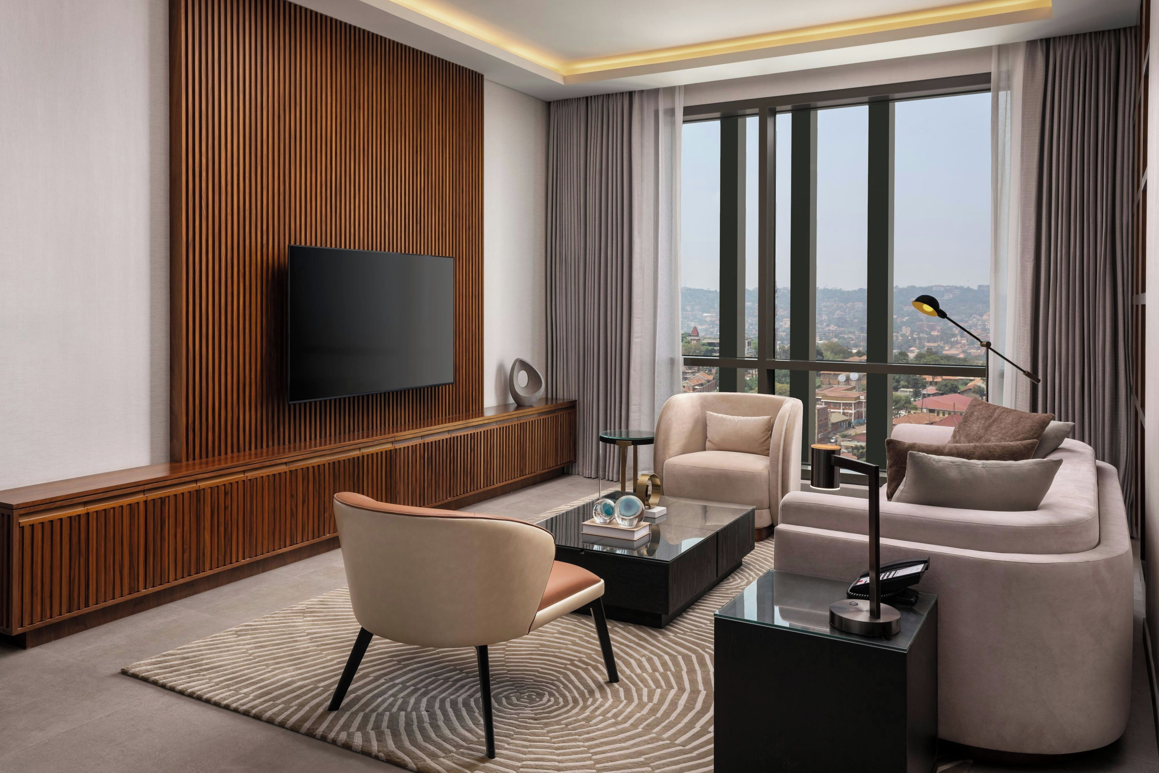 A modern living room with a flat-screen TV mounted on a wooden panel wall, a sofa, a coffee table, and a view of a cityscape through large windows.