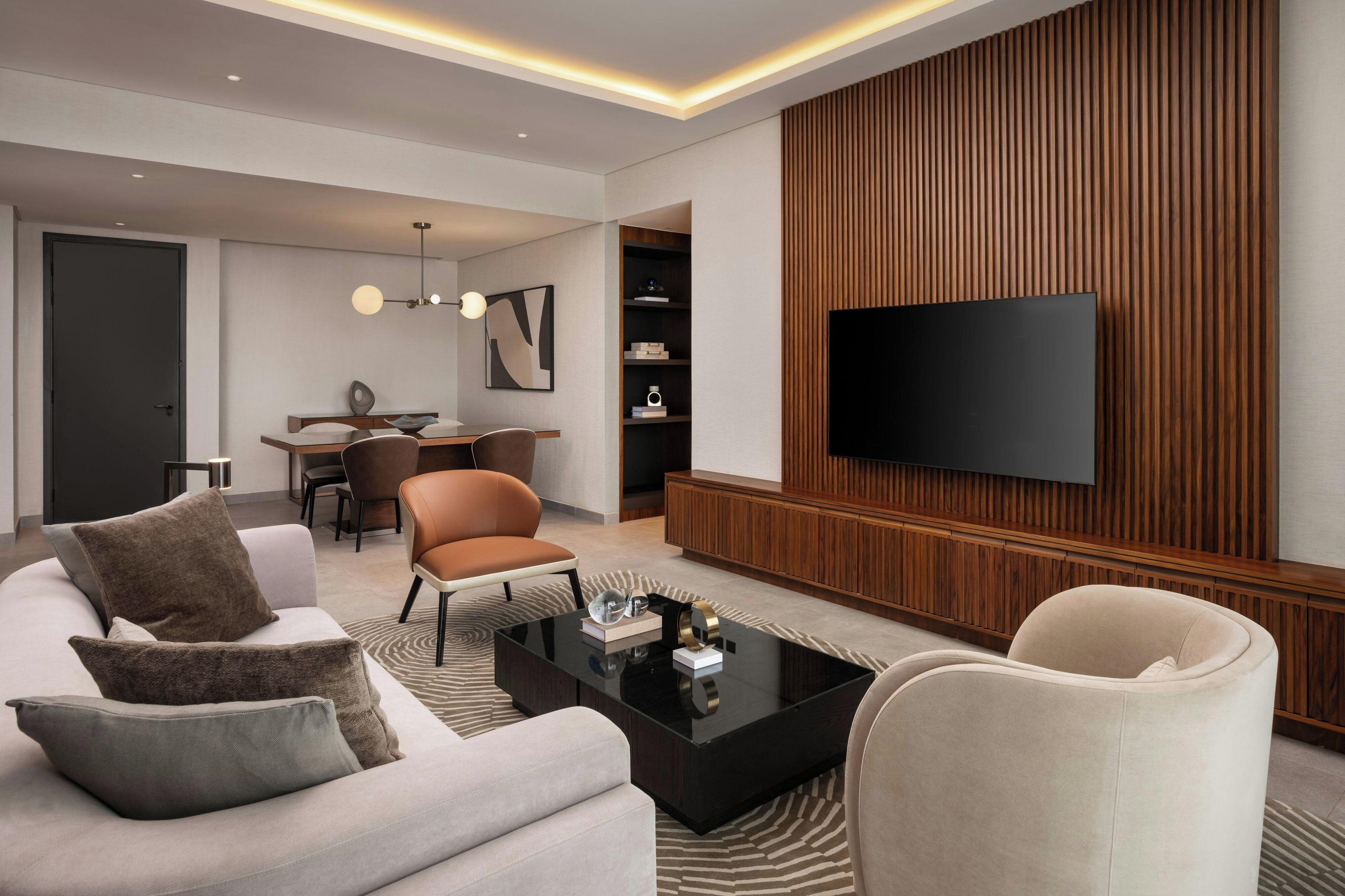 A modern living room with a sofa, armchairs, a coffee table, a flat-screen TV mounted on a wooden panel wall, and a dining area with a table and chairs.