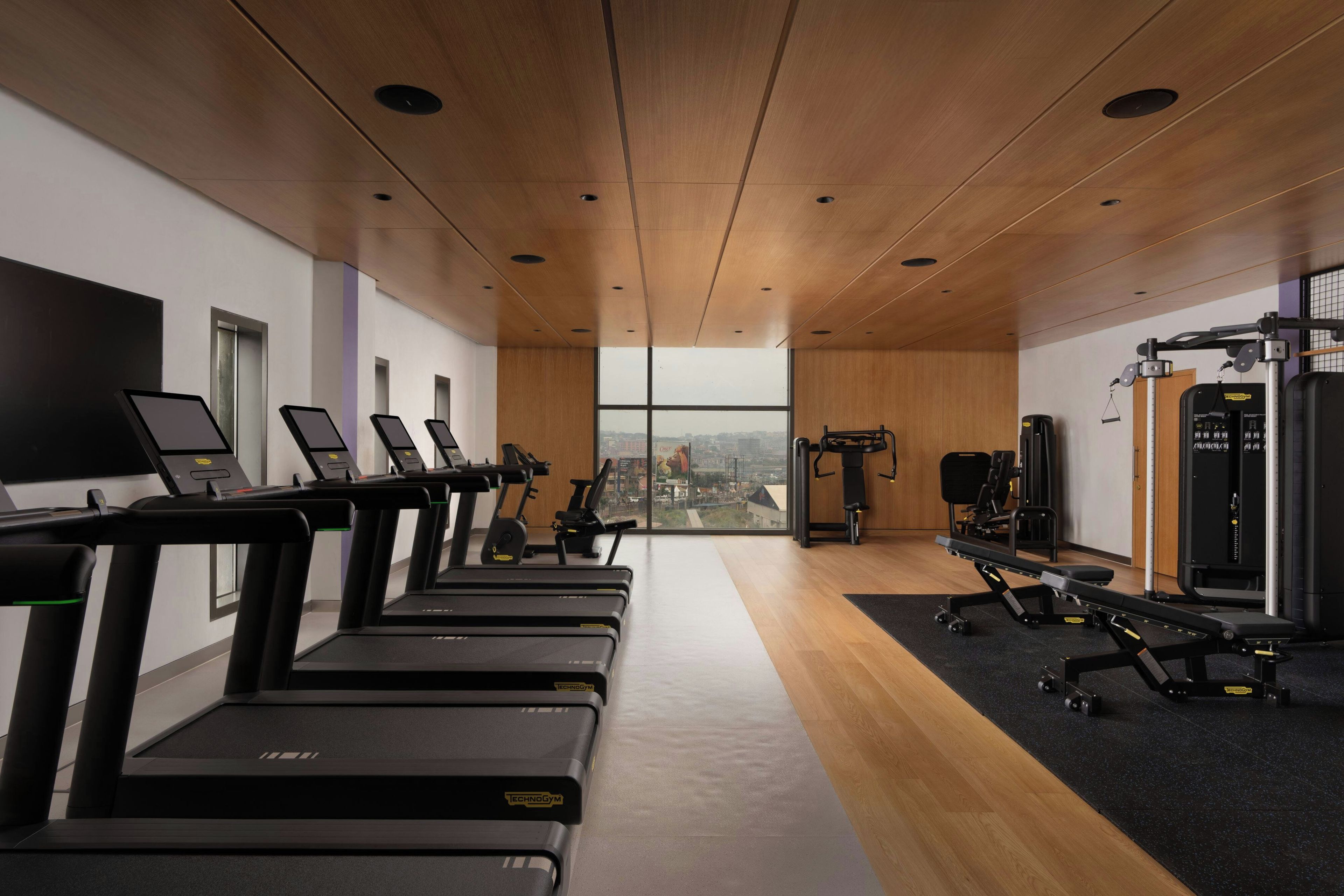 A gym with treadmills, weight machines, and a large window offering a city view.