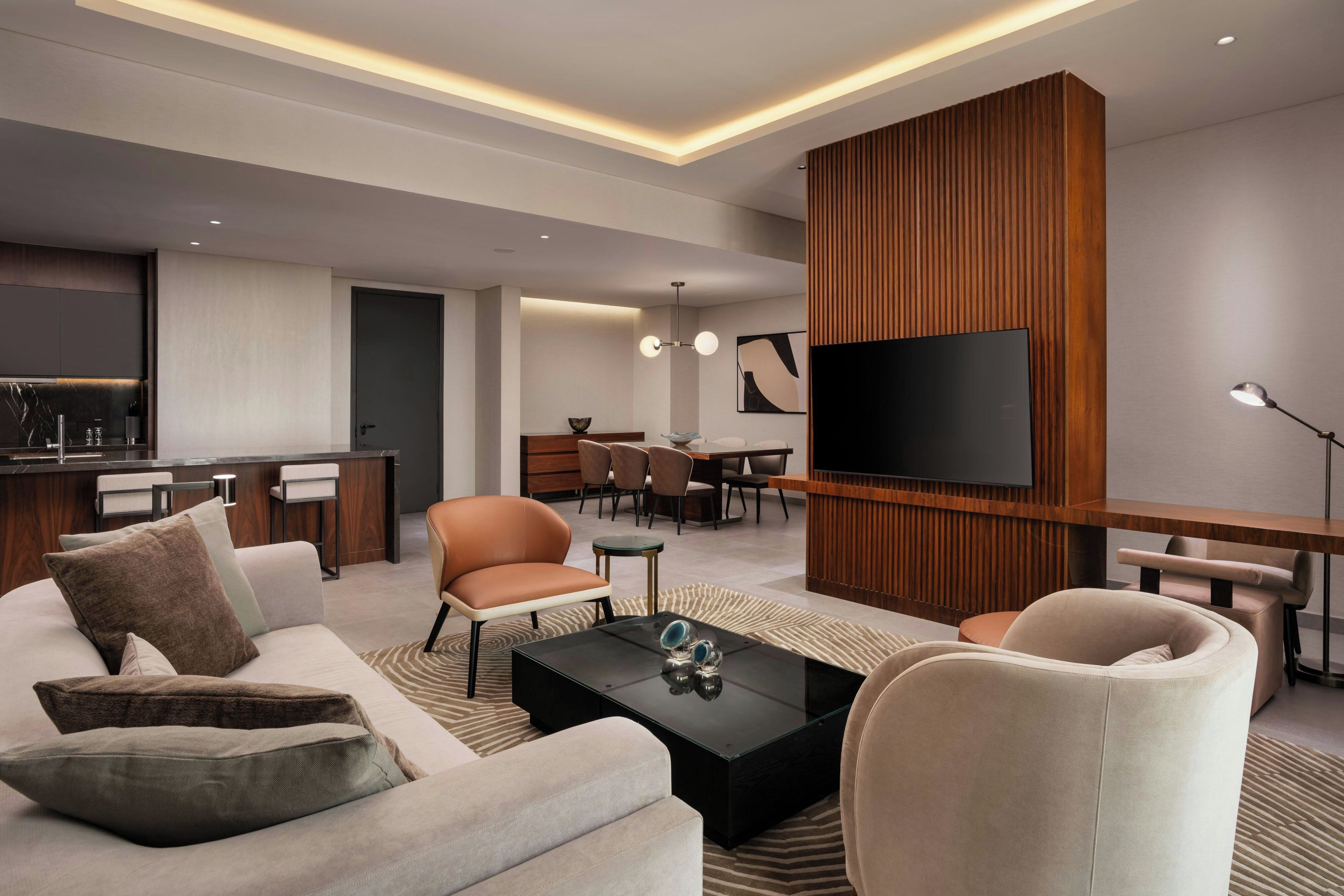 Ein modernes Wohnzimmer mit einer Couch, Sesseln, einem Couchtisch, einem Fernseher und einer Küchenzeile im Hintergrund.