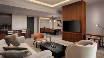Ein modernes Wohnzimmer mit einer Couch, Sesseln, einem Couchtisch, einem Fernseher und einer Küchenzeile im Hintergrund.