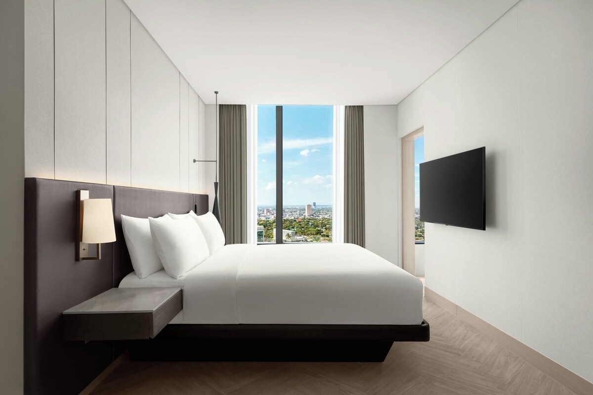 A modern hotel room with a bed, a bedside table, a TV mounted on the wall, and a large window offering a city view.