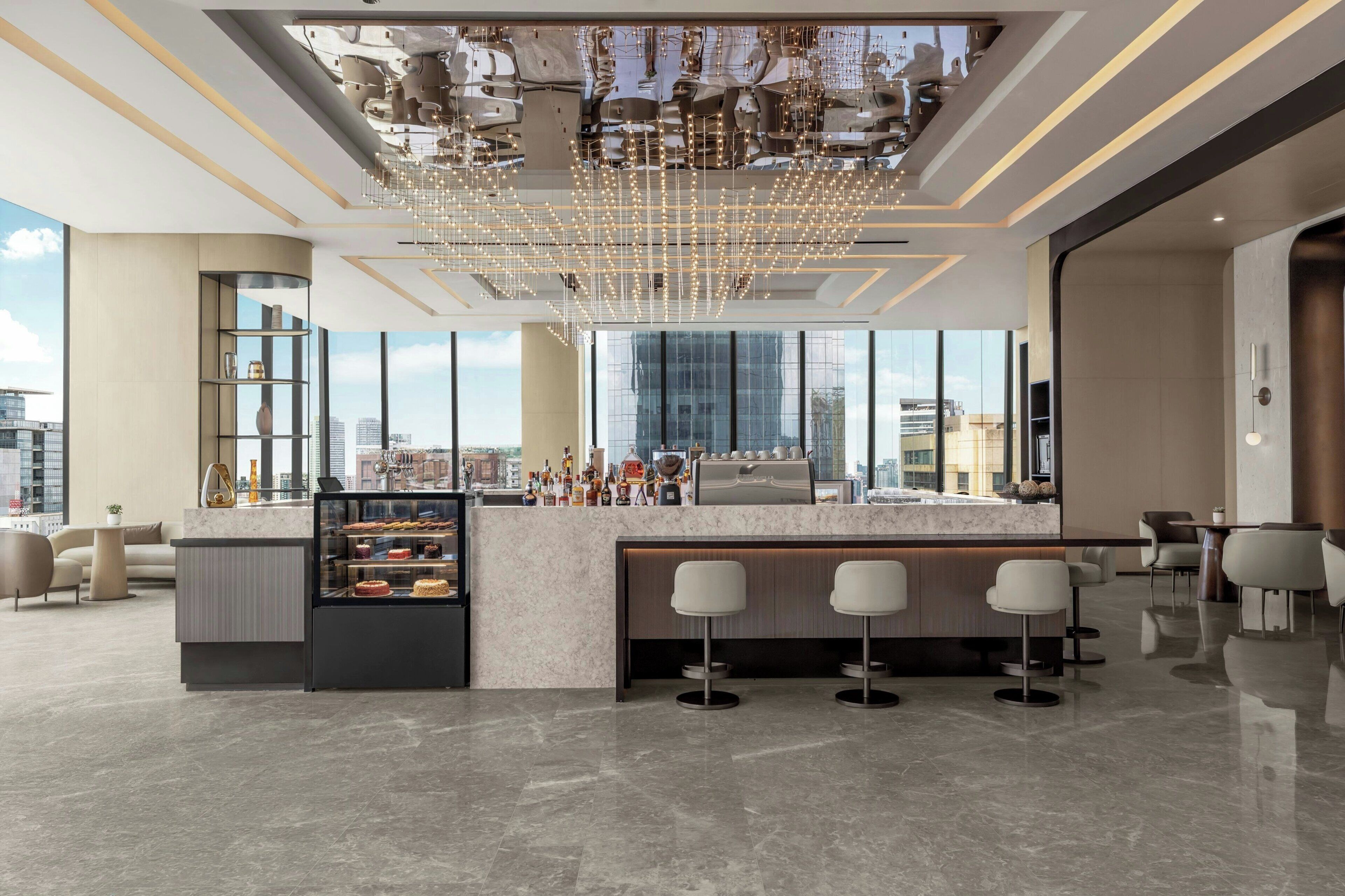 Zona de bar moderna con encimera de mármol, una vitrina con postres y vistas a la ciudad a través de grandes ventanales.