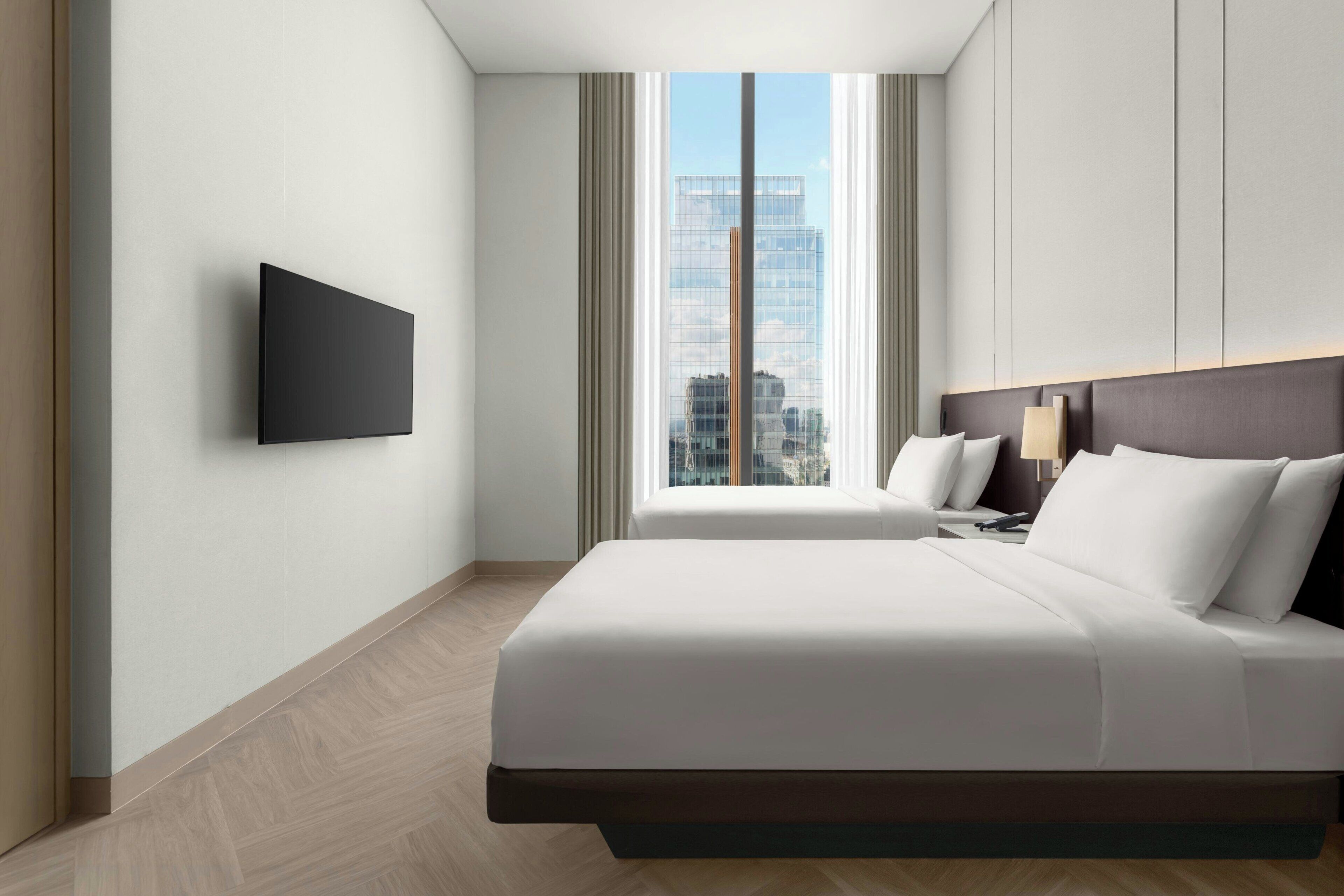 A hotel room with a bed, a flat-screen TV mounted on the wall, a window with curtains, and a city view.