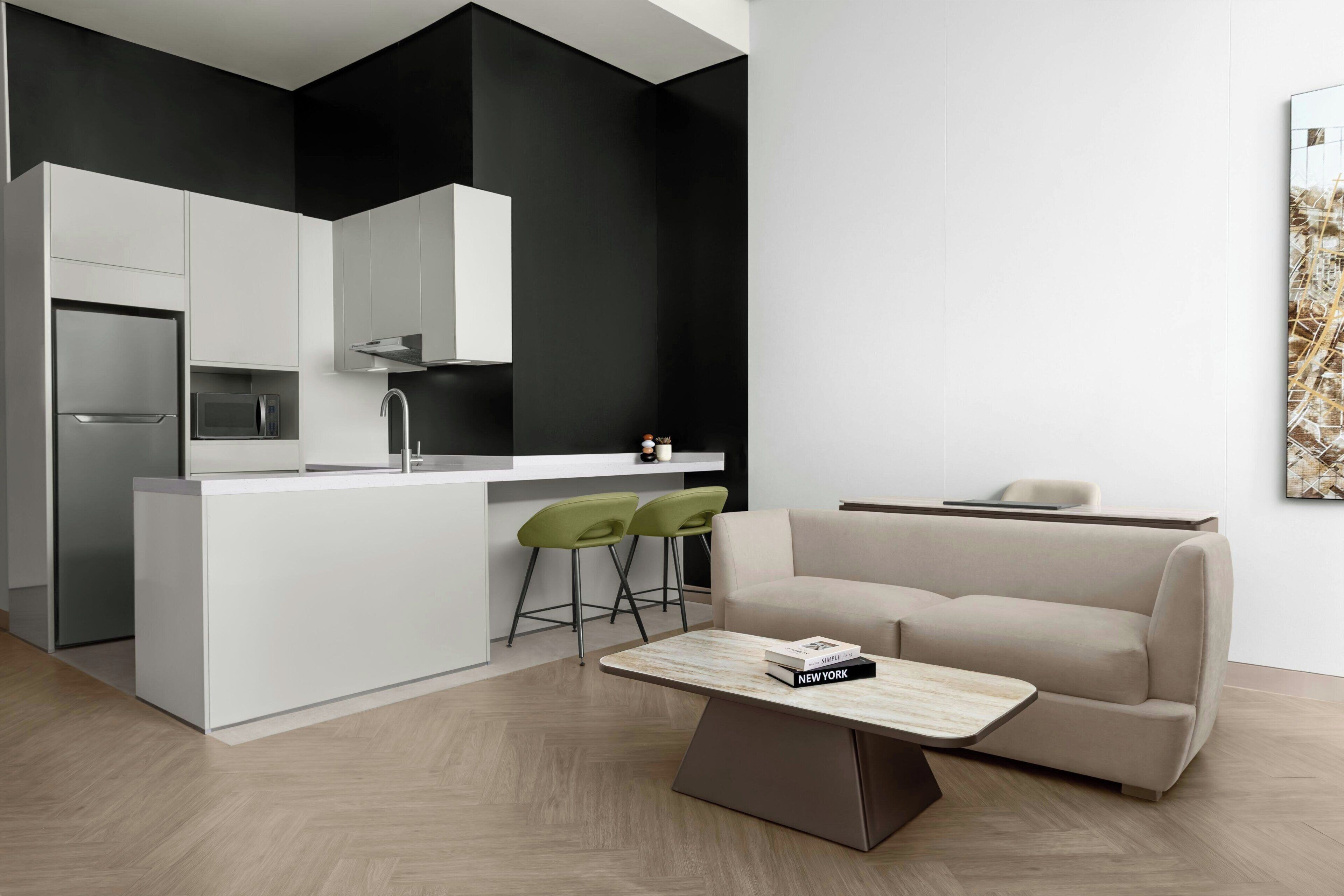 A modern kitchen with white cabinets, a stainless steel refrigerator, and a microwave. There is a dining area with a beige sofa and two green bar stools. A book titled 'Simple Living' is on the coffee table.