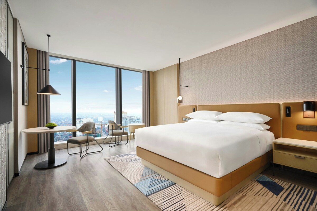 A modern hotel room with a large bed, a desk, a chair, and a city view.