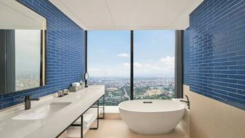 A modern bathroom with a large bathtub, dual sinks, and a city view through large windows.