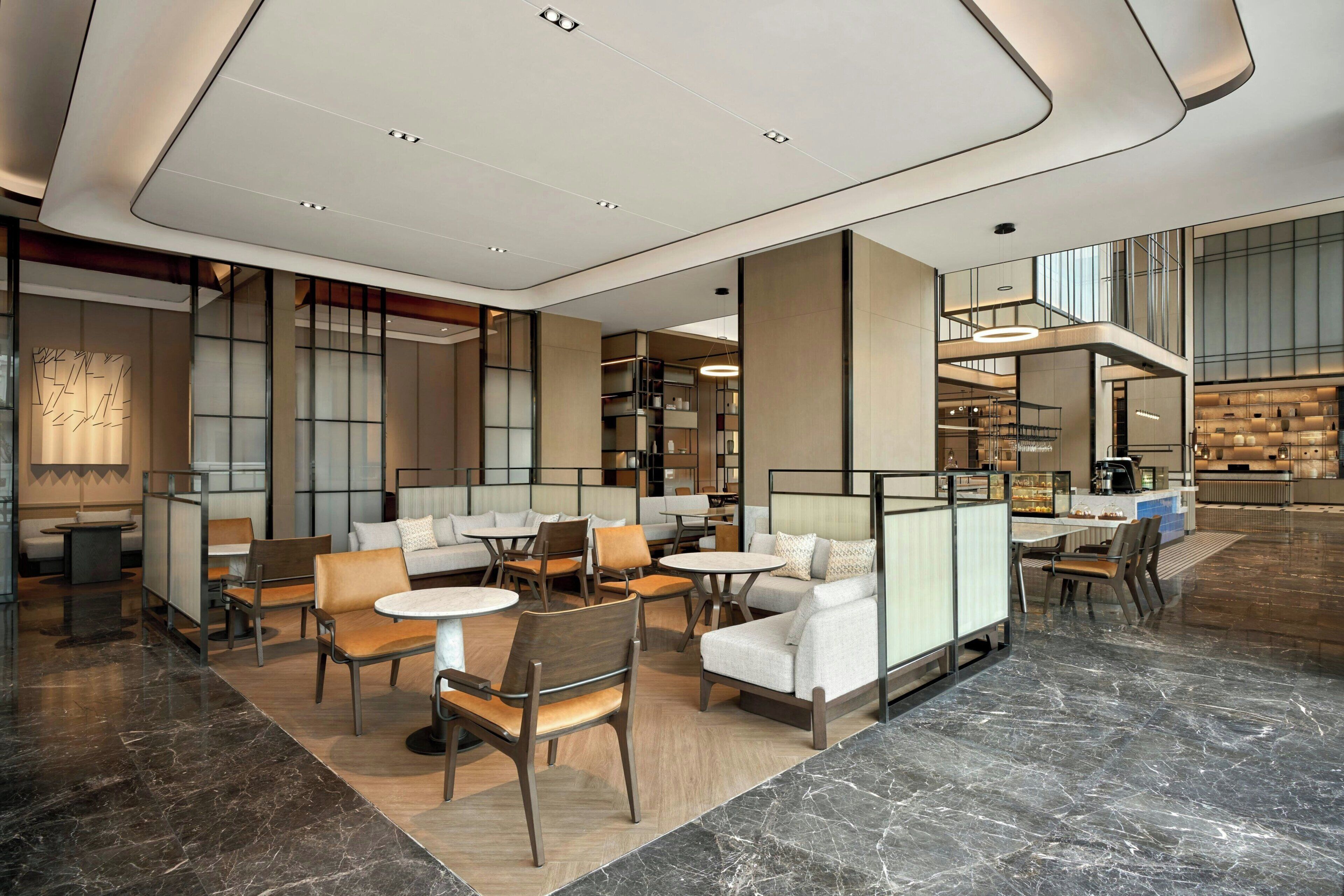 A modern hotel lobby with seating areas, a reception desk, and a view of the kitchen.