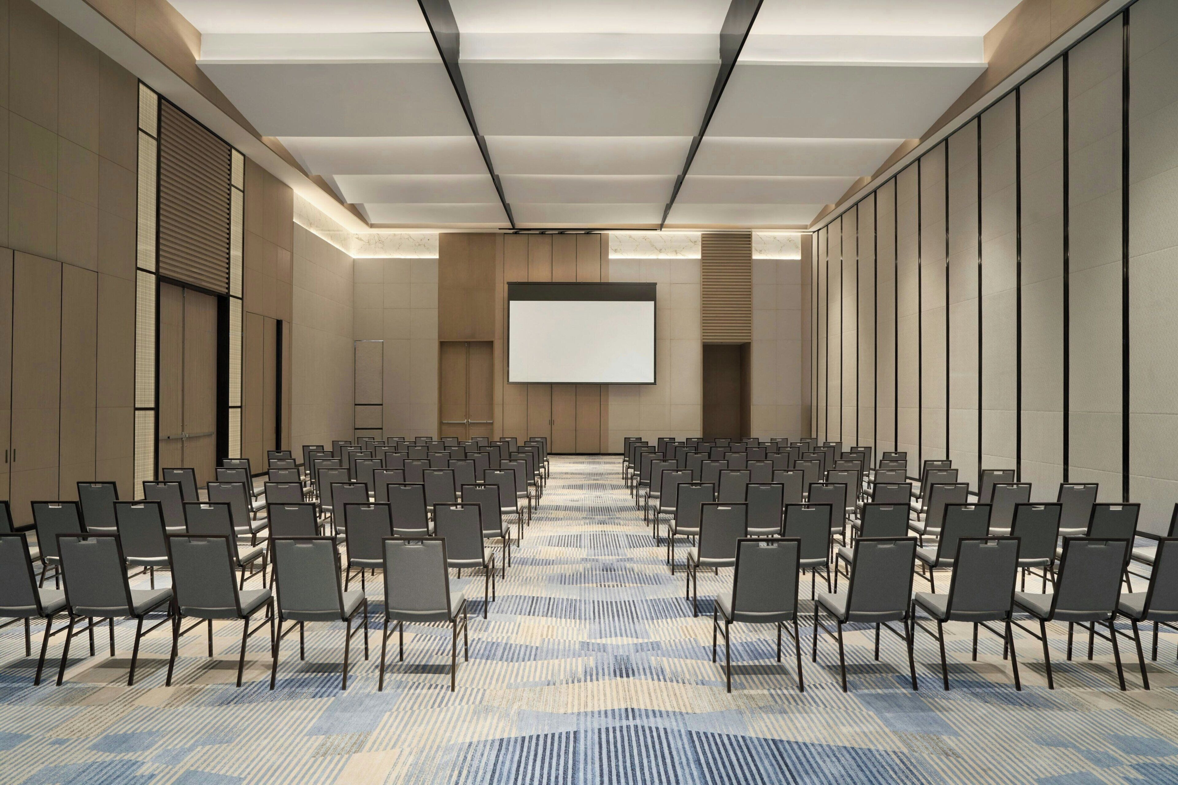 A conference room with rows of chairs, a large screen, and a carpeted floor.
