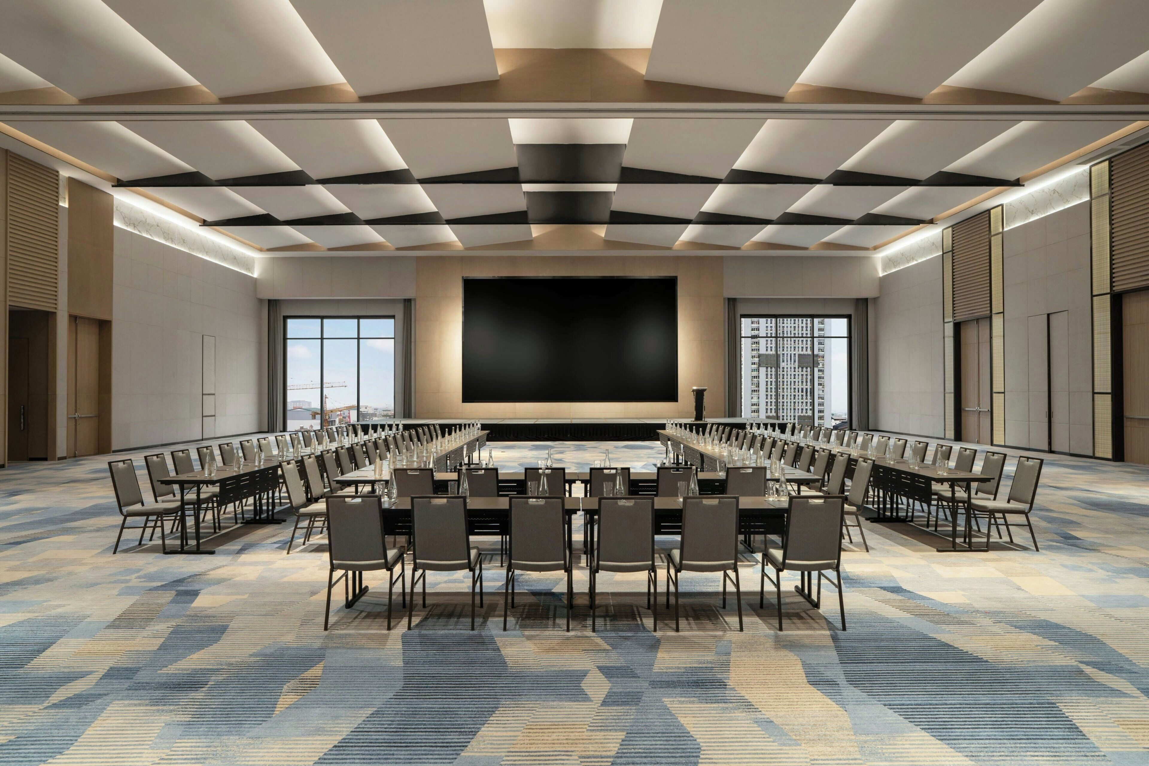 Un salón de conferencias amplio con un escenario central, varias filas de sillas y grandes ventanales que ofrecen vistas a la ciudad.