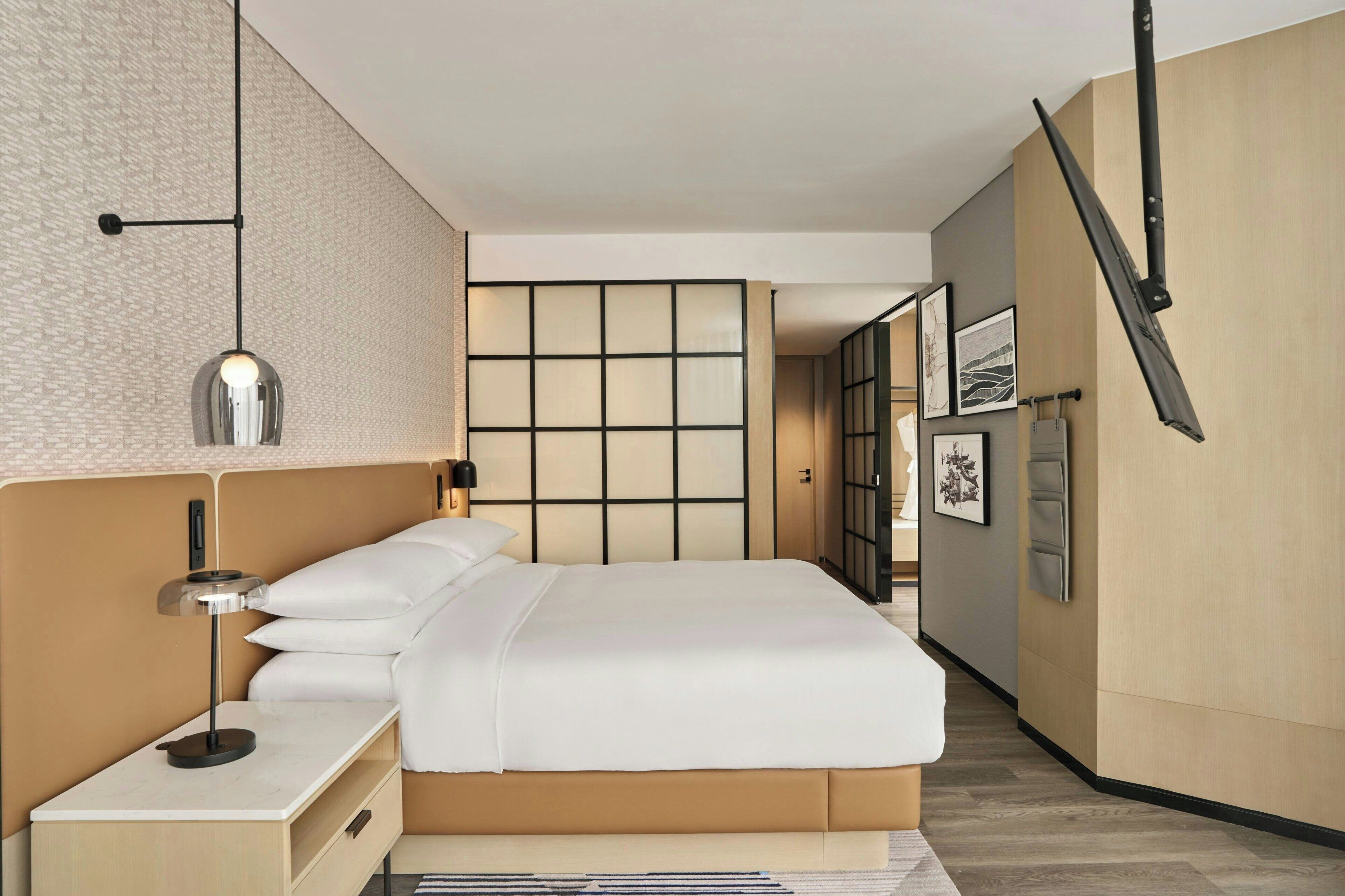 A modern hotel room with a large bed, a bedside table with a lamp, a wall-mounted TV, and a sliding door leading to another room.