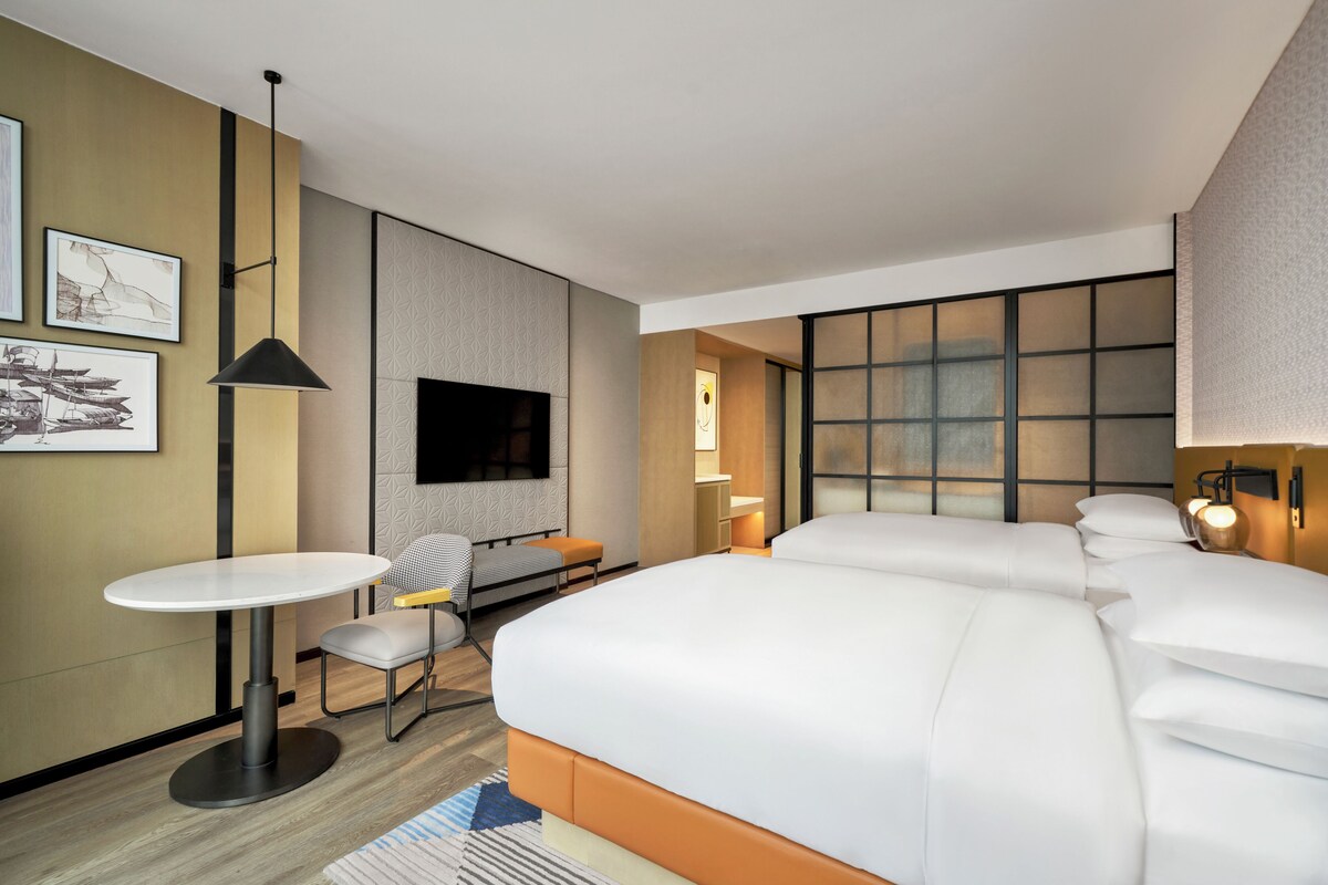 A hotel room with a bed, a desk with a chair, a TV, and a large window.