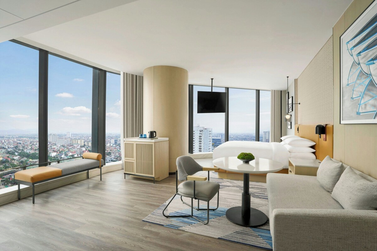 A modern hotel room with a large window, a sofa, a dining table, a chair, a small cabinet, and a TV mounted on the wall.