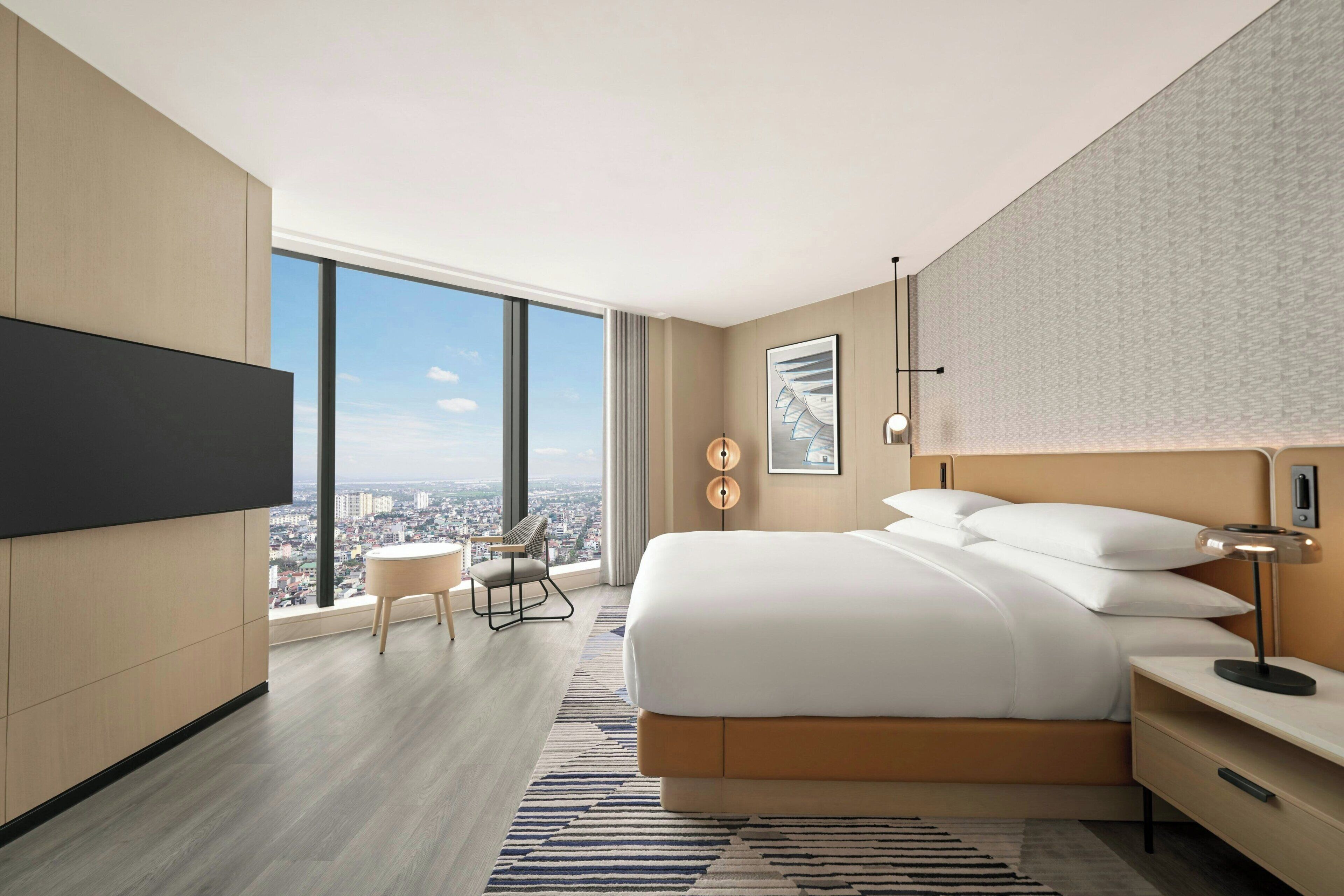 Una habitación de hotel moderna con una cama grande, un escritorio, una silla, un televisor y vistas a la ciudad.