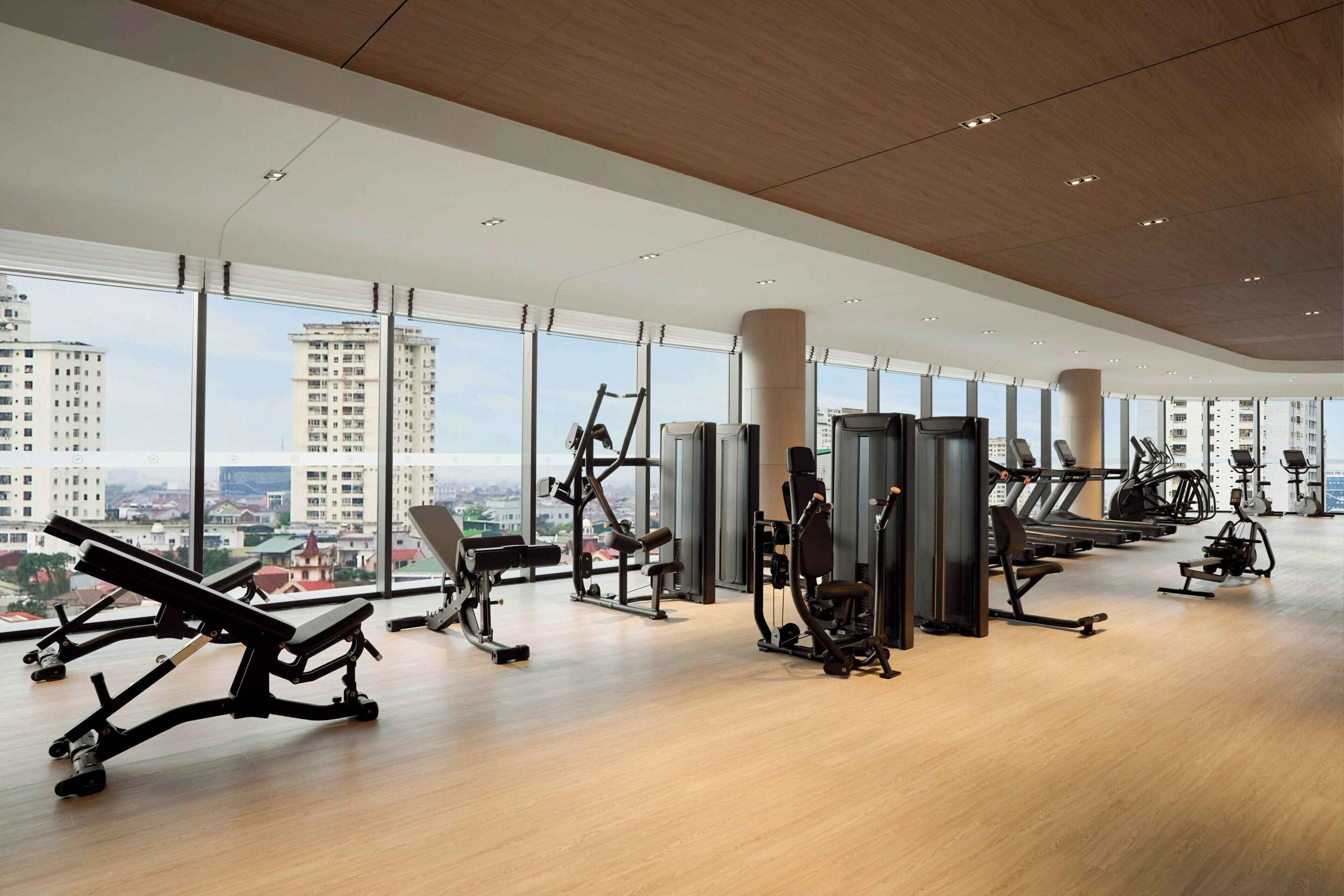 A modern gym with various exercise equipment, large windows, and a city view.