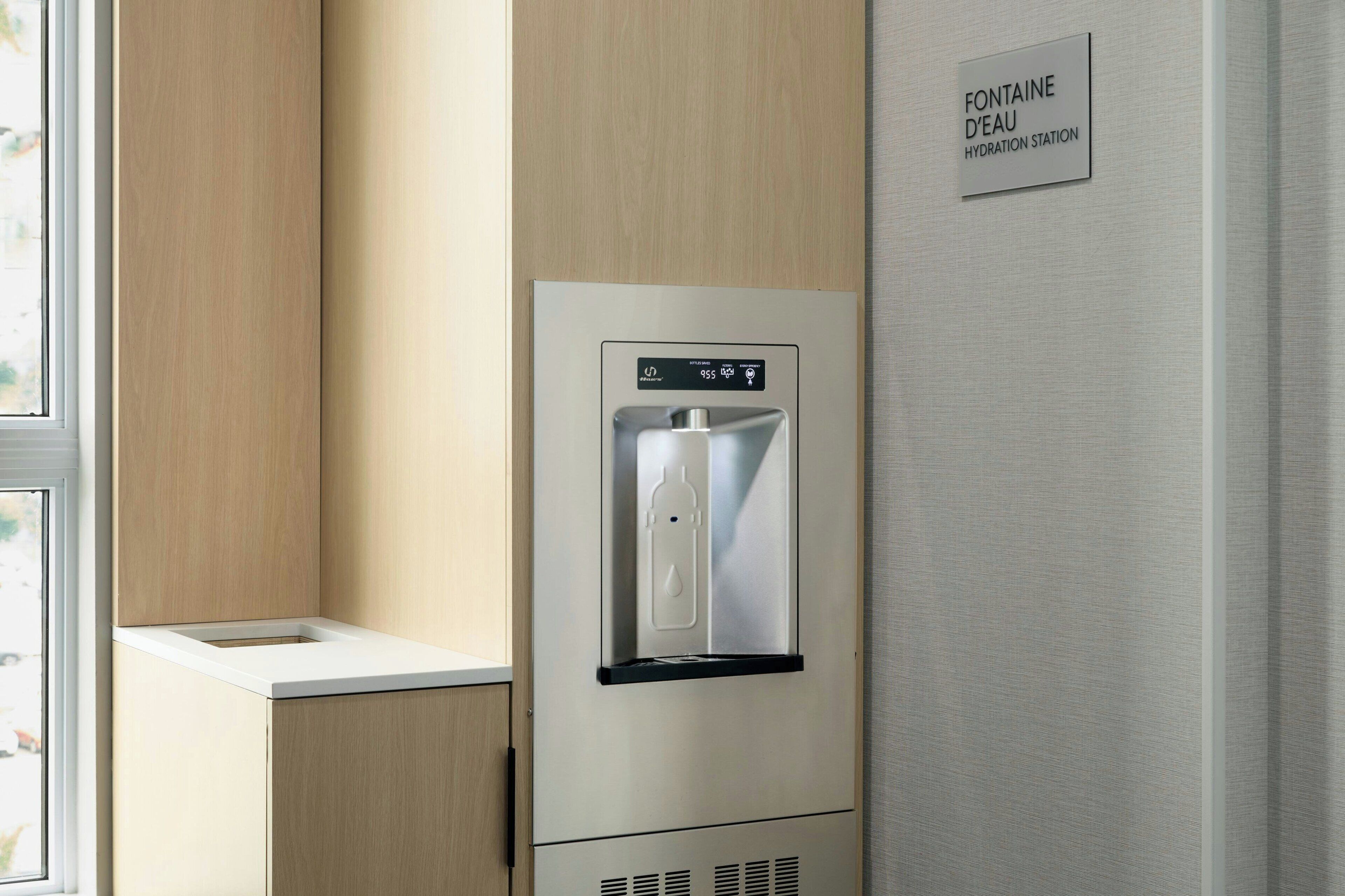 Ein Wasserspender mit digitaler Anzeige, platziert in einem Raum mit Holzwandverkleidung und einem Schild, das ihn als Trinkstation kennzeichnet.