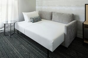 Ein Zimmer mit einem Einzelbett, einem grauen Sofa, einem kleinen runden Tisch und einem gemusterten Zierkissen.