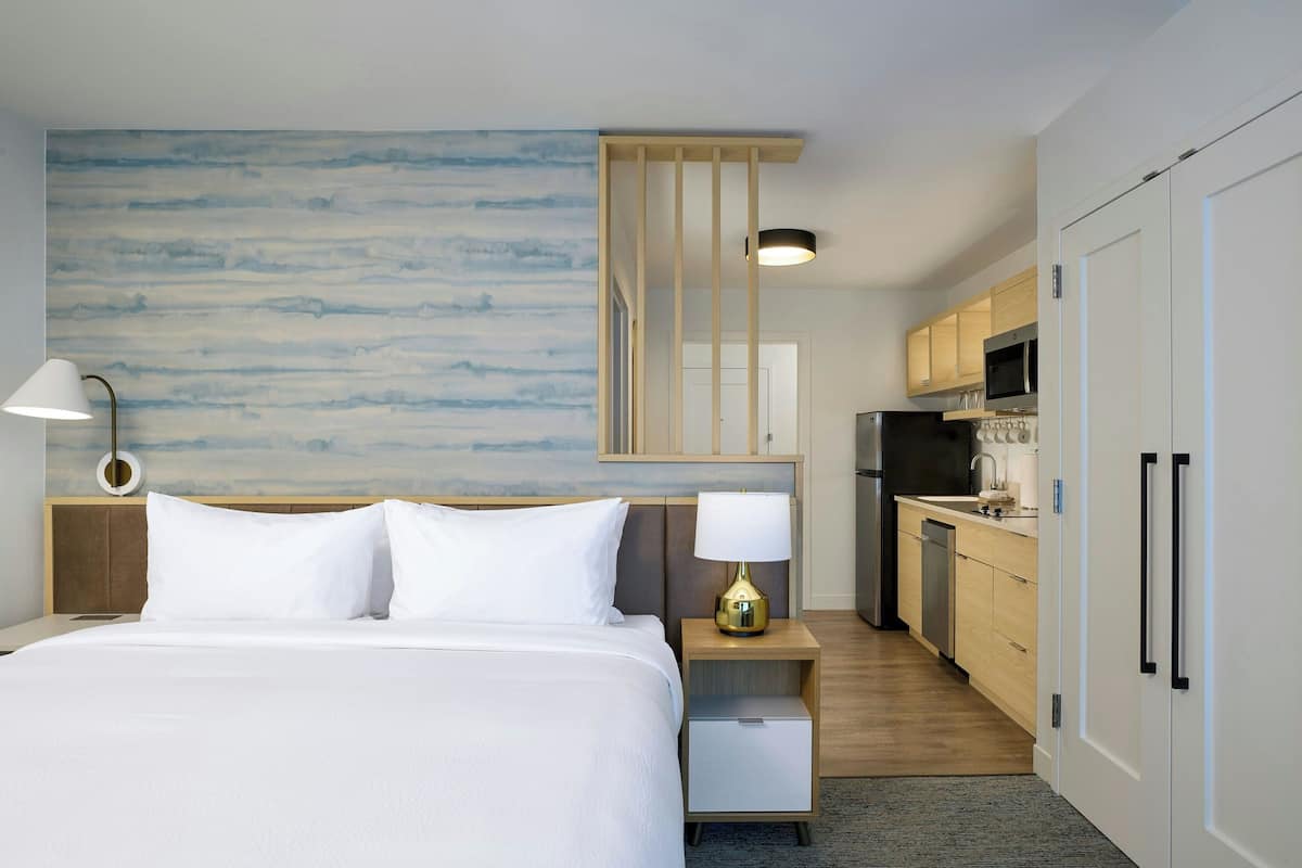 Una habitación de hotel con una cama, una lámpara de noche, una mesita de noche y vistas a la cocina.