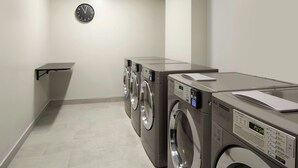 A laundry room with two front-loading washing machines, a wall-mounted clock, and a small shelf. - Towneplace Suites By Marriott Montreal Airport (Dorval)