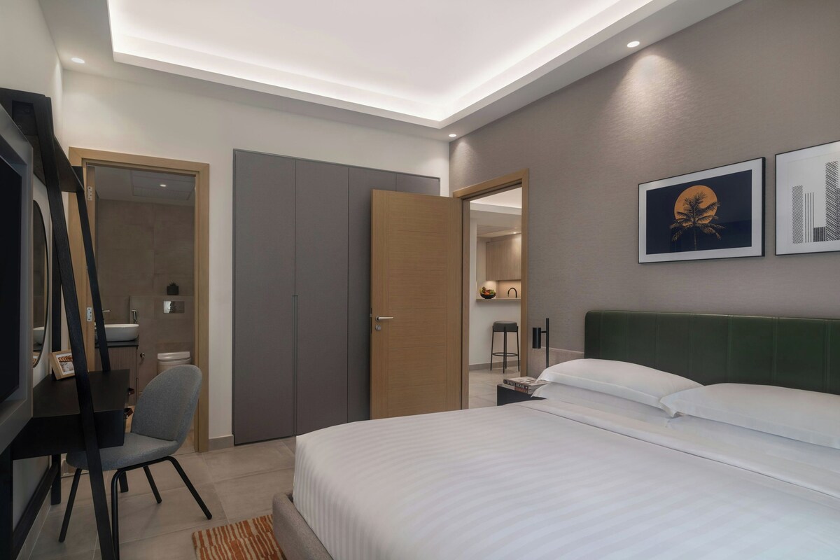 Habitación de hotel moderna con cama, escritorio, silla, armario y baño visible a través de una puerta abierta.