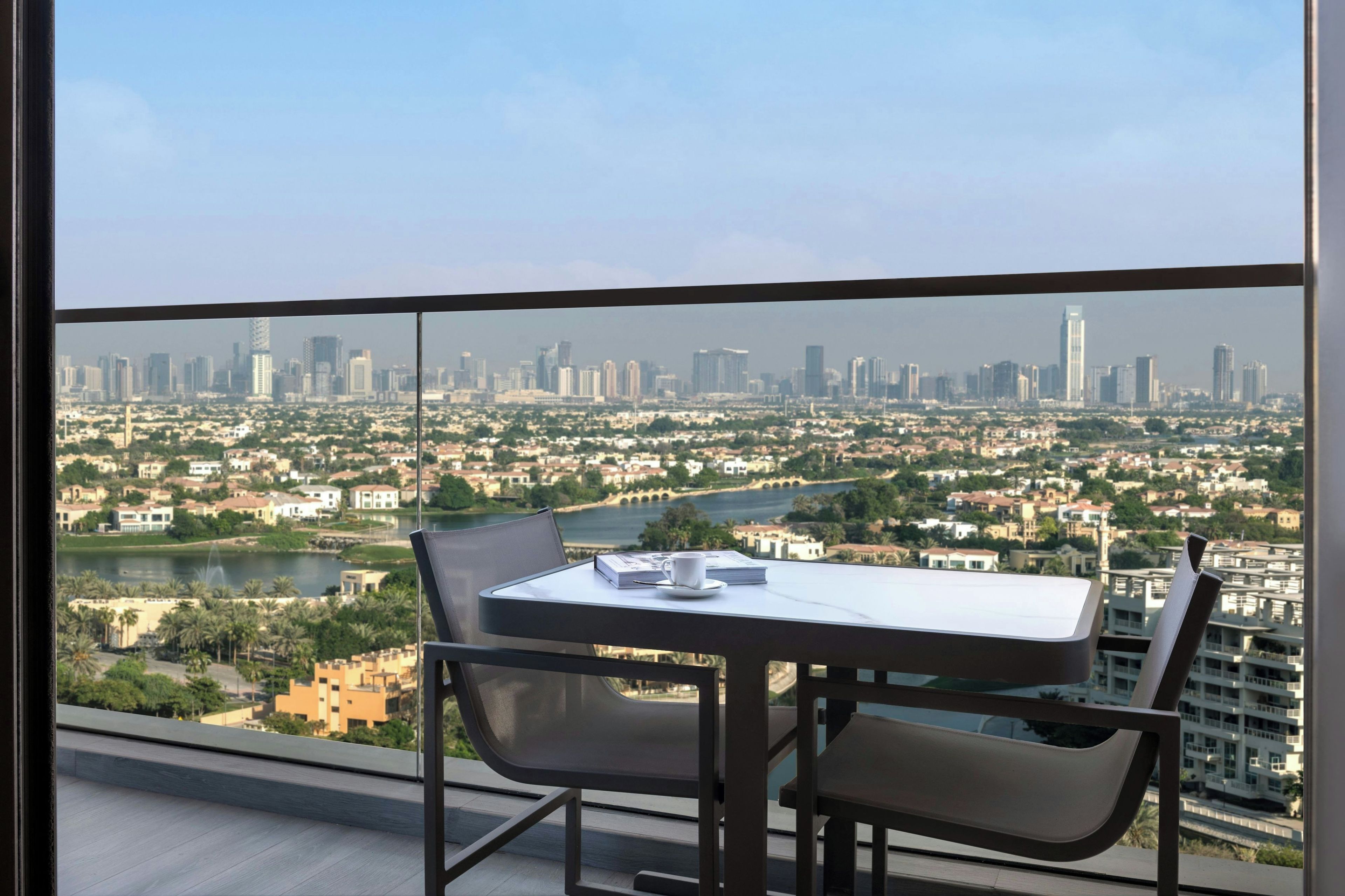 A balcony with a table and chairs, overlooking a cityscape with buildings and a river.