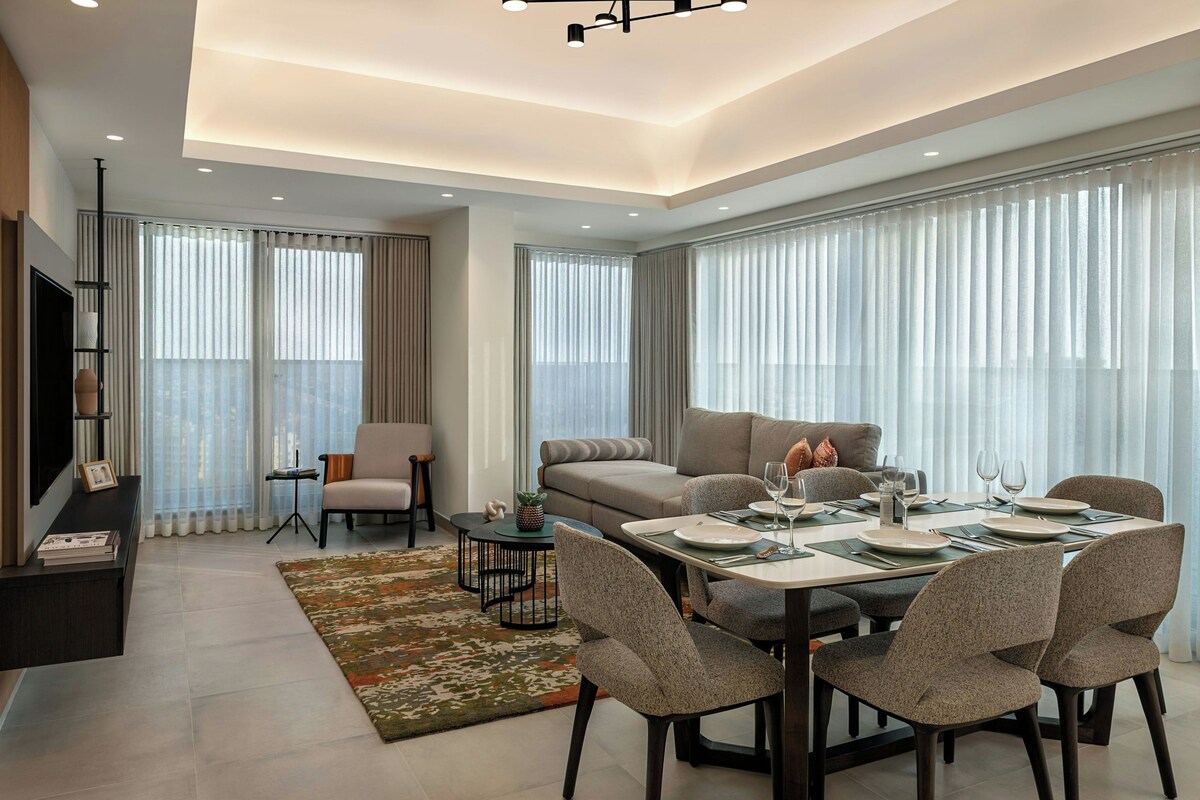 Una sala de estar moderna con comedor, ventanas amplias con cortinas, un televisor de pantalla plana y una mesa de centro.