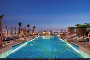 A rooftop infinity pool with a city view, surrounded by lounge areas and palm trees.