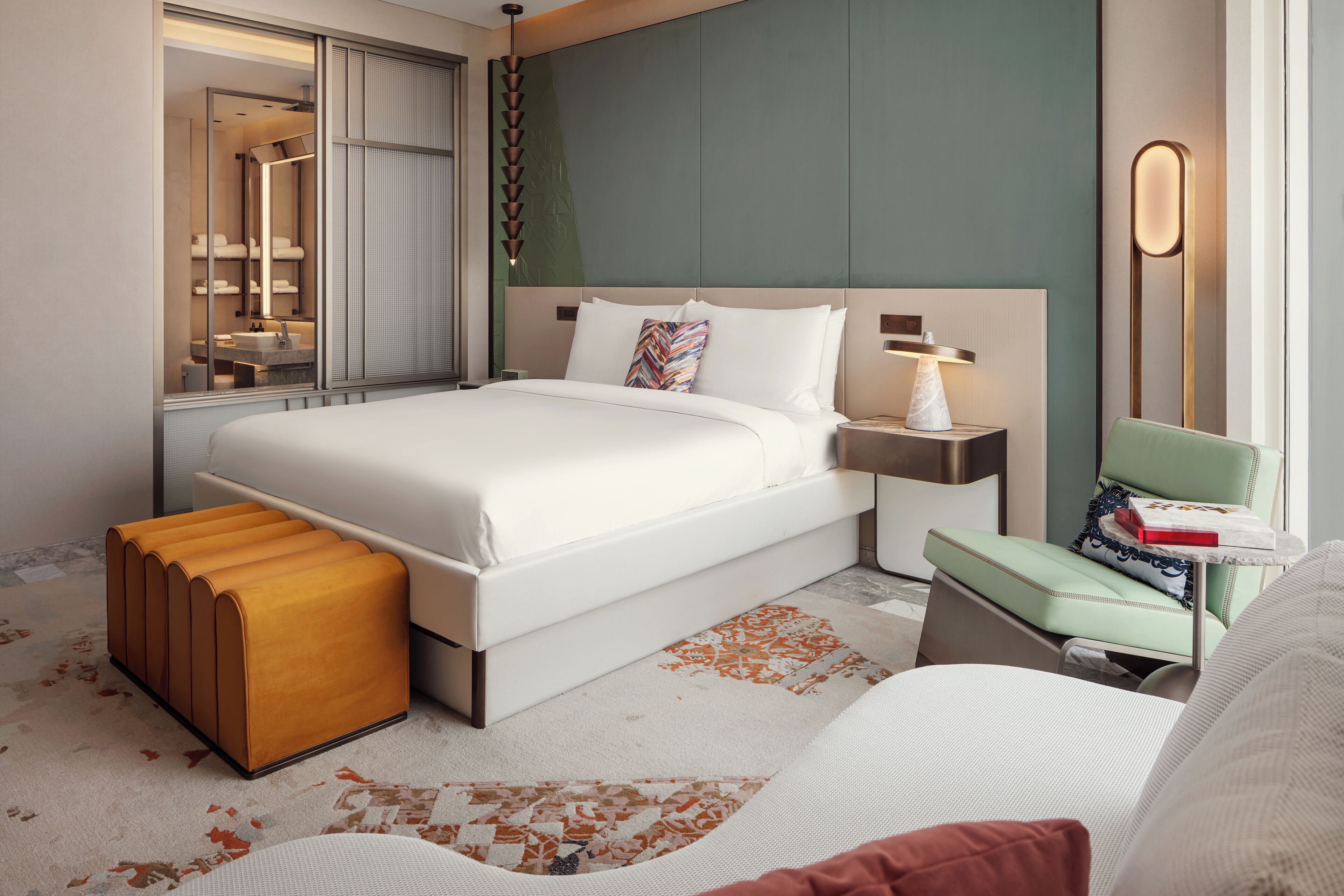 Une chambre d’hôtel moderne avec un grand lit, une table de chevet, une commode, un banc, un miroir et une vue sur la salle de bain.