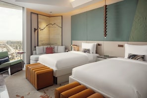 A hotel room with two beds, a large window with a city view, a decorative wall piece, and a hanging light fixture.