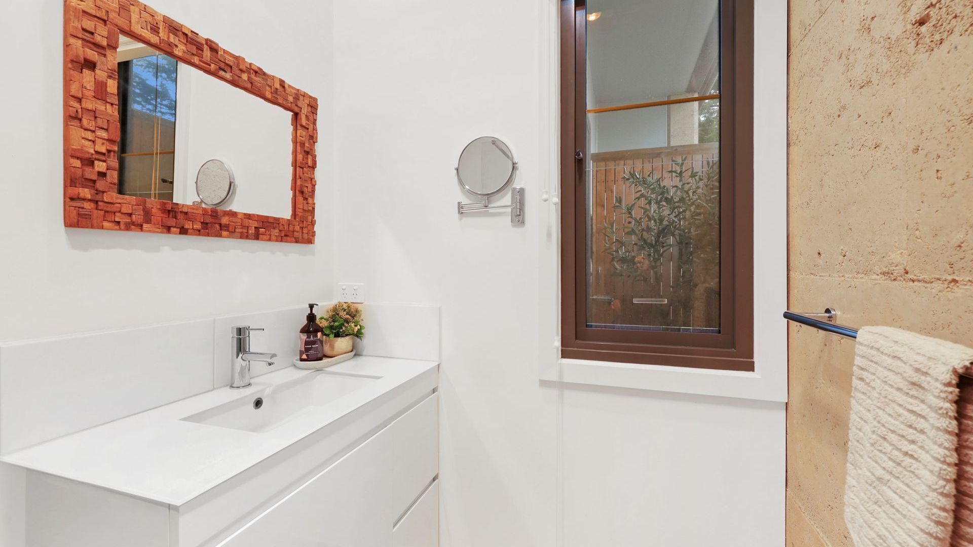 A bathroom with a white vanity, a mirror with a brick frame, a round mirror, a window with a view of greenery, and a towel rack.