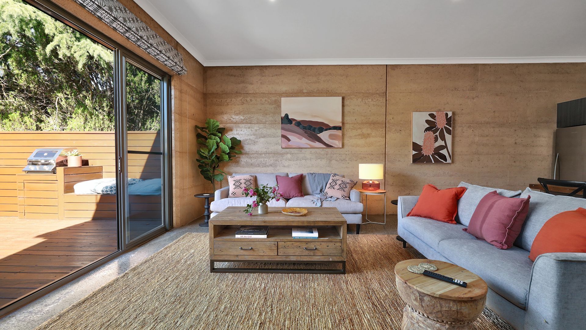 A modern living room with a wooden coffee table, a grey sofa, and a white sofa with colorful cushions. There is a painting on the wall and a plant on the floor.