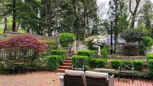 Un patio con mobili in vimini, pavimento in mattoni, un giardino con diverse piante e una recinzione in legno.
