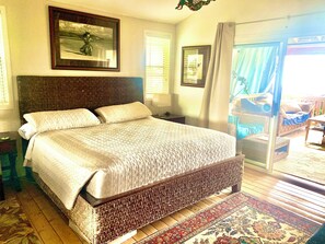 A bedroom with a large bed, a wooden headboard, a nightstand, a bedside table, a window with blinds, and a view of an outdoor balcony area. - Peaceful 3-bedroom bungalow in enchanting Kailua-Kona perfect for relaxing (Kailua-Kona)