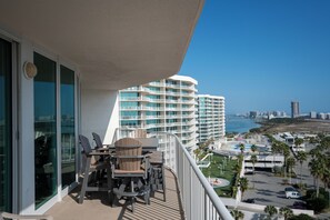 Uma varanda com mesa e cadeiras, vista para uma praia com edifícios altos e um céu azul límpido.